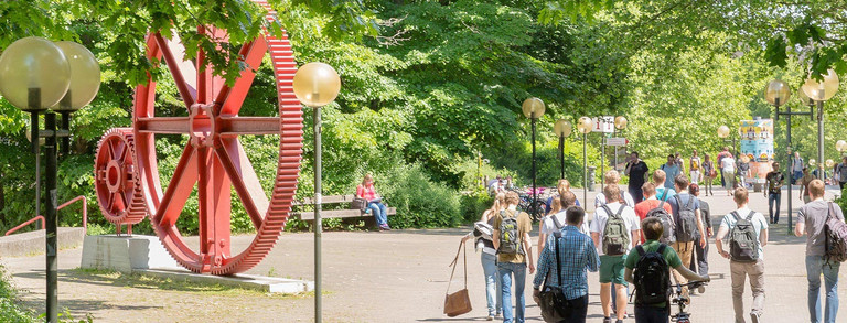 Header image students red gears campus north Students in summer on north campus, red gears in background.